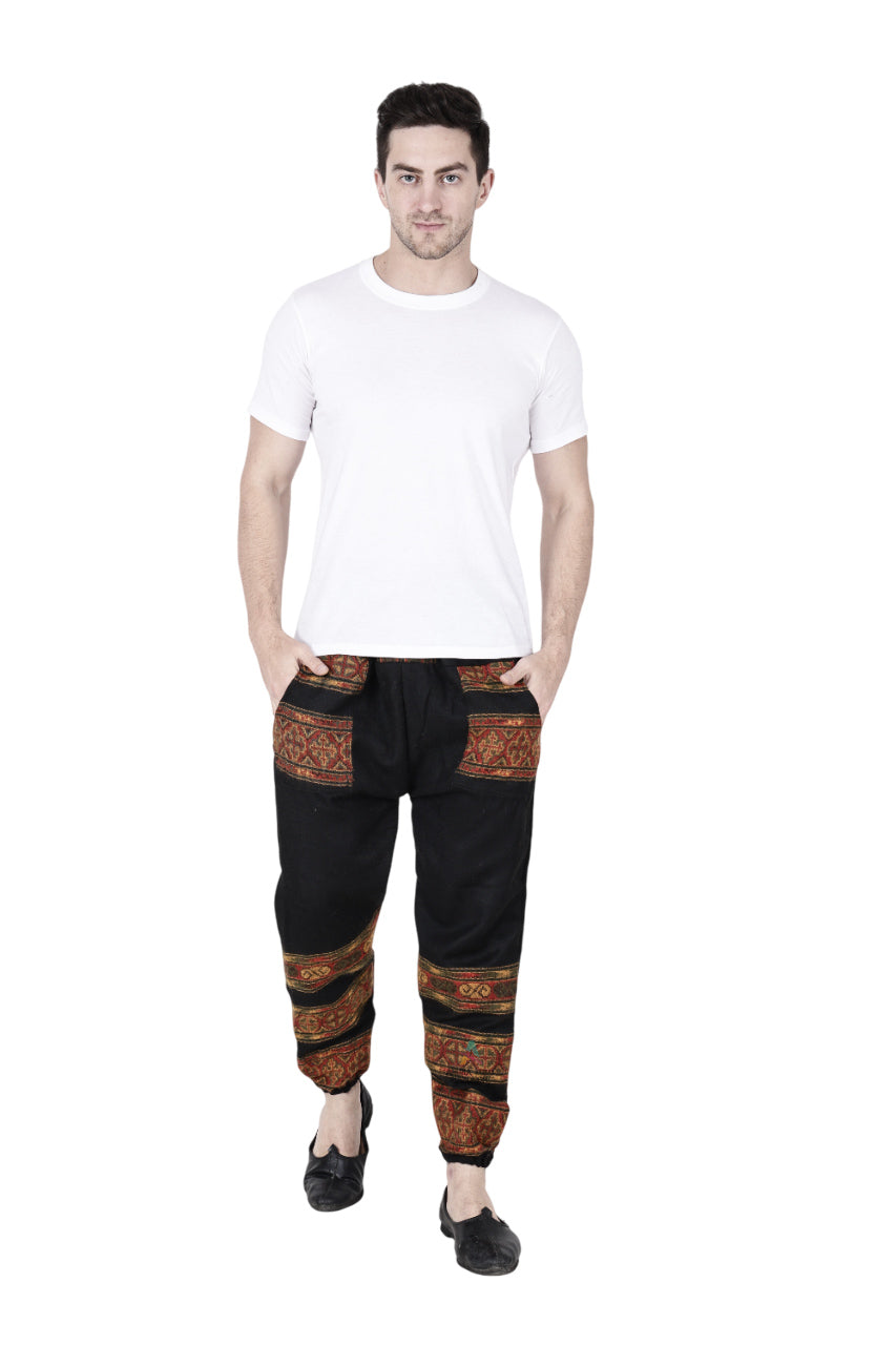 A man wears aruna kullu handloom's woolen pajamas for winter in black, featuring red and orange patterned bands, paired with a plain white t-shirt and black shoes, standing with hands in pockets against a white background.