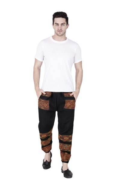 A man wears aruna kullu handloom's woolen pajamas for winter in black, featuring red and orange patterned bands, paired with a plain white t-shirt and black shoes, standing with hands in pockets against a white background.