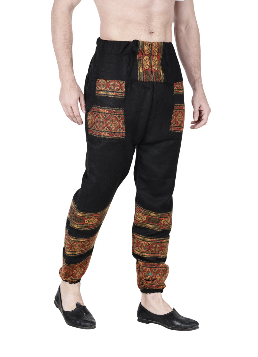A person wearing aruna kullu handloom Woolen pajamas for winter black, featuring colorful embroidery on the pockets, waistband, and legs, paired with black slip-on shoes, stands against a white background.