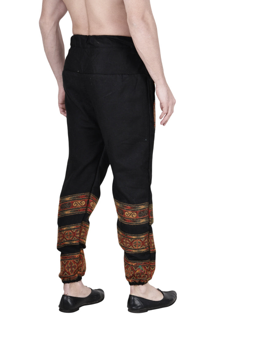 A person stands barefoot, facing away, wearing aruna kullu handloom woolen pajamas for winter in black with colorful traditional patterns on the lower legs and no shirt, set against a white background.