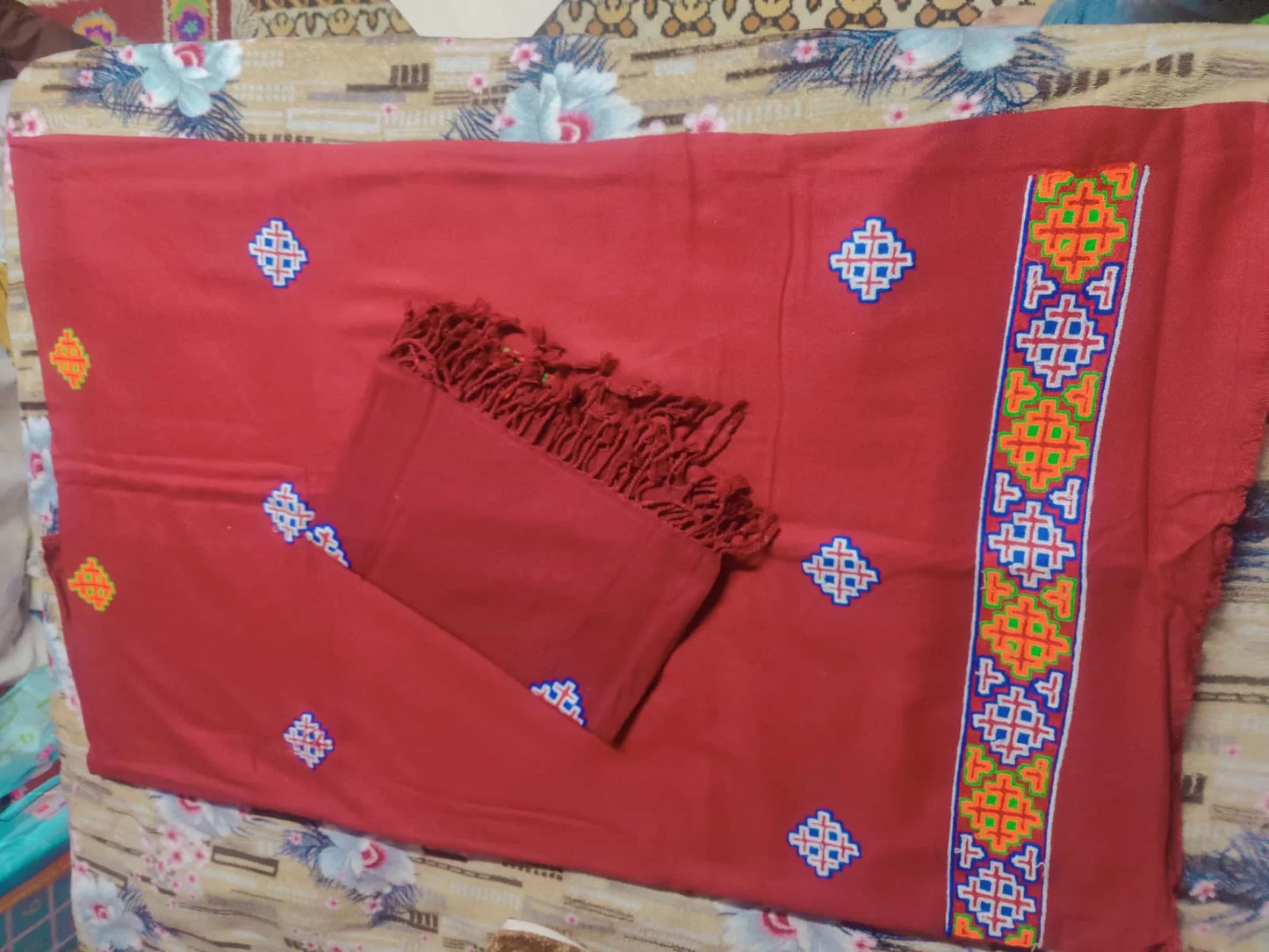 Aruna Kullu Handloom's "Kullu Patti Woolen Suit 4 Meter with Stole" in maroon features traditional geometric embroidery in blue, yellow, red, and green motifs along the edge; includes a matching fringed stole and is set against a floral bedsheet.
