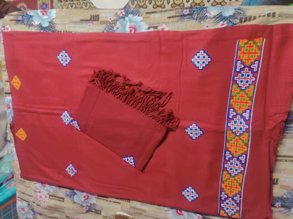Aruna Kullu Handloom's "Kullu Patti Woolen Suit 4 Meter with Stole" in maroon features traditional geometric embroidery in blue, yellow, red, and green motifs along the edge; includes a matching fringed stole and is set against a floral bedsheet.