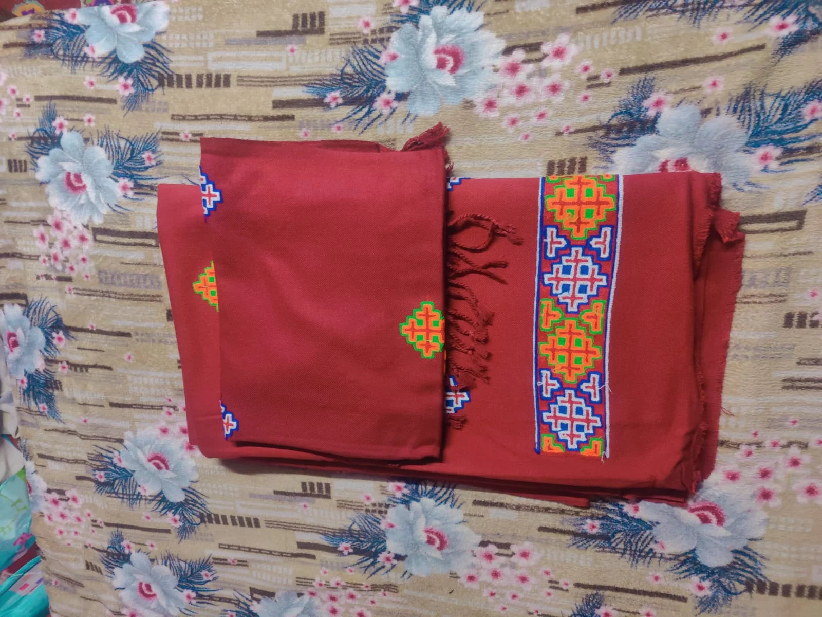 The aruna kullu handloom kullu patti woolen suit (4 meter) with stole in maroon features colorful geometric embroidery in yellow, blue, green, and red on folded fabric, displayed on a floral-patterned beige surface.