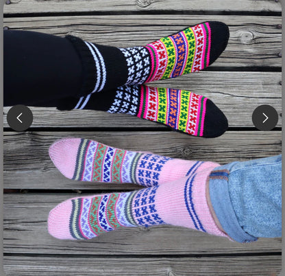 Two pairs of feet in aruna kullu handloom Woollen Traditional Hand Made Kullu Socks (Multicolour, Free Size) pink rest on a wooden floor—one pair features black striped socks, the other showcases colorful pink ladies' designs.