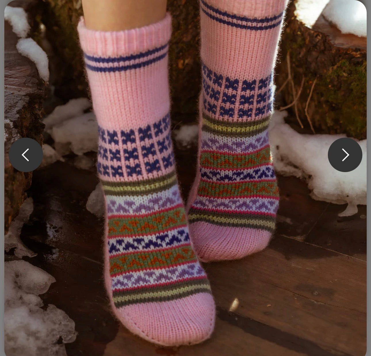 A person sports aruna kullu handloom's Woollen Traditional Hand Made Kullu Socks (Multicolour, Free Size, pink), featuring cozy and vibrant patterns, while standing on a wooden surface near snow and tree trunks.