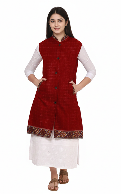 A woman faces forward wearing the aruna kullu handloom Check red long jacket with kullu lace work over a white dress. She has dark hair, hands in pockets, brown sandals, and stands against a plain white background.