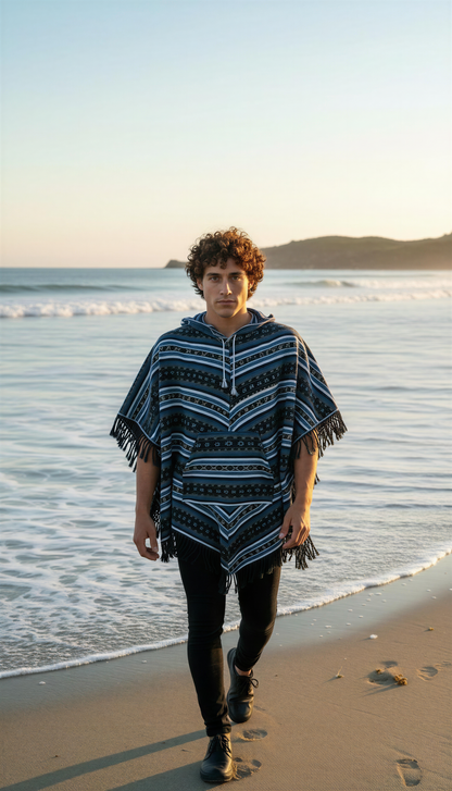 Wearing the aruna kullu handloom Hippie poncho blue, a curly-haired person walks barefoot along the shoreline at sunset, with gentle waves and distant hills in the background.