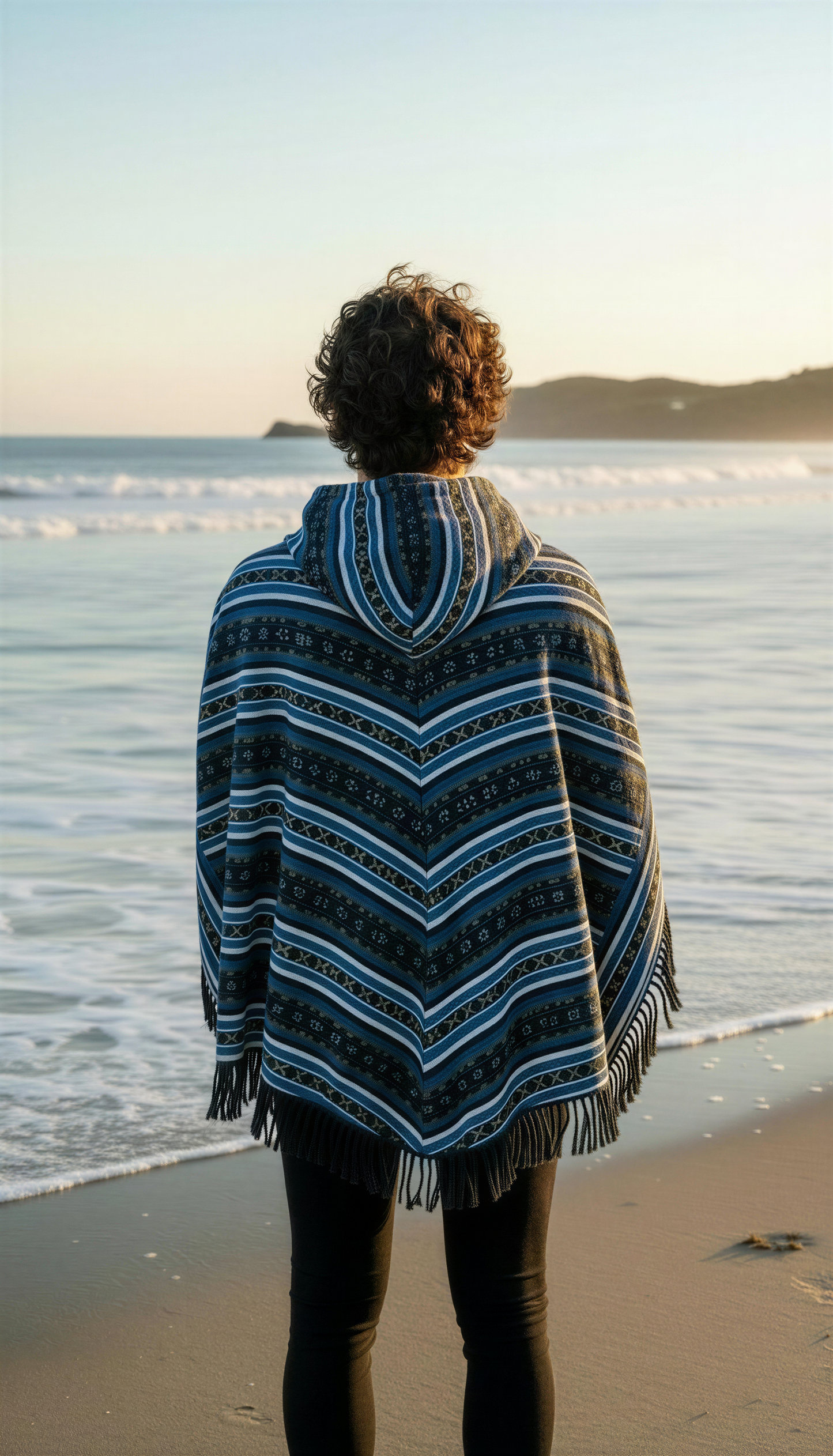 A person with curly hair wearing the Hippie poncho blue by aruna kullu handloom stands on the shore at sunset, facing the ocean as gentle waves roll in and distant hills form the backdrop.