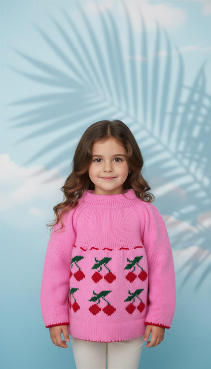 A young girl with long brown hair wears the aruna kullu handloom PURE WOOL HAND KNITTED woolen baby set in pink with a cherry design, standing before a light blue background with palm leaf shadow patterns.