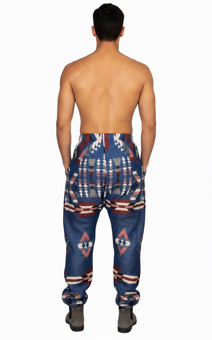 A shirtless man faces away, wearing aruna kullu handloom Wool Super Soft Warm Pajamas WITH KULLU PATTI WORK in blue, featuring bold geometric and tribal-inspired patterns in white, red, and beige. He also wears gray shoes against a plain white background.