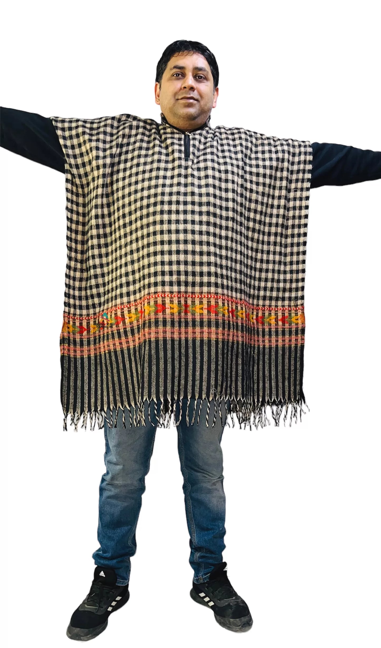 A man stands with arms outstretched, wearing the aruna kullu handloom "kullu shawl woolen kaftan for men check black," featuring a black and white check design, colorful stripe near the bottom, and fringe hem. Background is plain white.