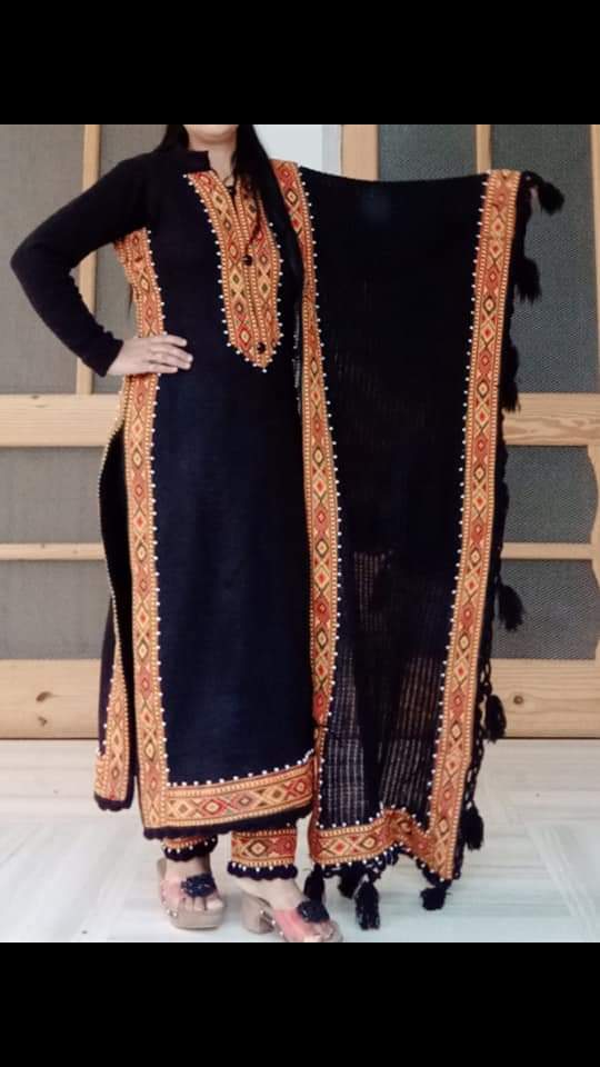 A woman wears a suit adorned with aruna kullu handloom’s KULLU Patti (LACE) Border for Suits Kurtis and other dress material (3 M), paired with matching pants, a tasseled dupatta, and Pula Chappal sandals, standing indoors by wood and glass.