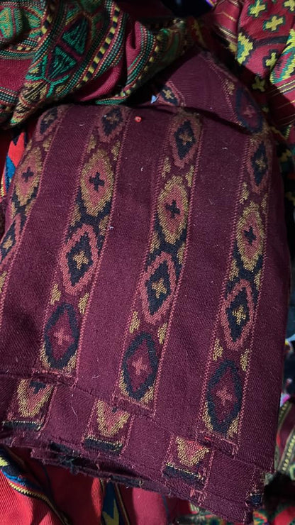 A close-up of aruna kullu handloom’s KULLU Patti (LACE) Border for Suits, Kurtis, and Dress Material (3 M) shows maroon fabric with black, gold, and red geometric patterns and a kullu border among other vibrant textiles.