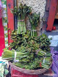 Bundles of fresh fiddlehead ferns for aruna kullu handloom’s Lingdu Ka Achar (लिंगड़ू का अचार | Fiddlehead Ferns Pickle | Kasrod Ka Achar) are stacked in a woven basket at an outdoor market, some tied and some loose, against a colorful backdrop.