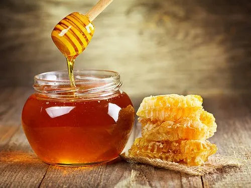 A glass jar of aruna kullu handloom Himachali Wild honey Raw honey with a honey dipper dripping honey sits on a wooden surface beside pieces of fresh honeycomb.