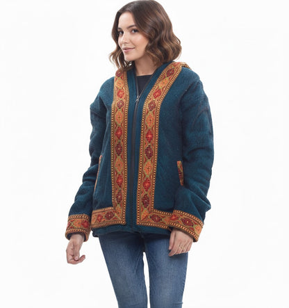 A woman with wavy brown hair wears the aruna kullu handloom Himachal handicrafts blue woolen winter zipper hoodie jacket for men & women, featuring colorful Kullu Patti work, paired with blue jeans against a white background.