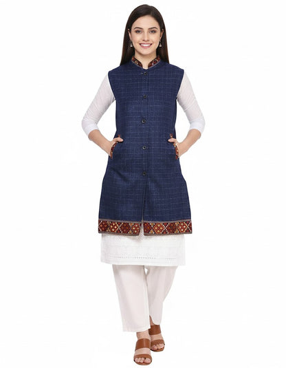 A woman smiles, posing in an aruna kullu handloom "Check navy blue long jacket with kullu lace work" over a white kurta and pants. She wears brown sandals and keeps her hands in the jacket's pockets against a white background.half jacket for women khadi half jacket for ladies ladies half jacket cotton women's half jacket ladies sadri design winter half jacket for women